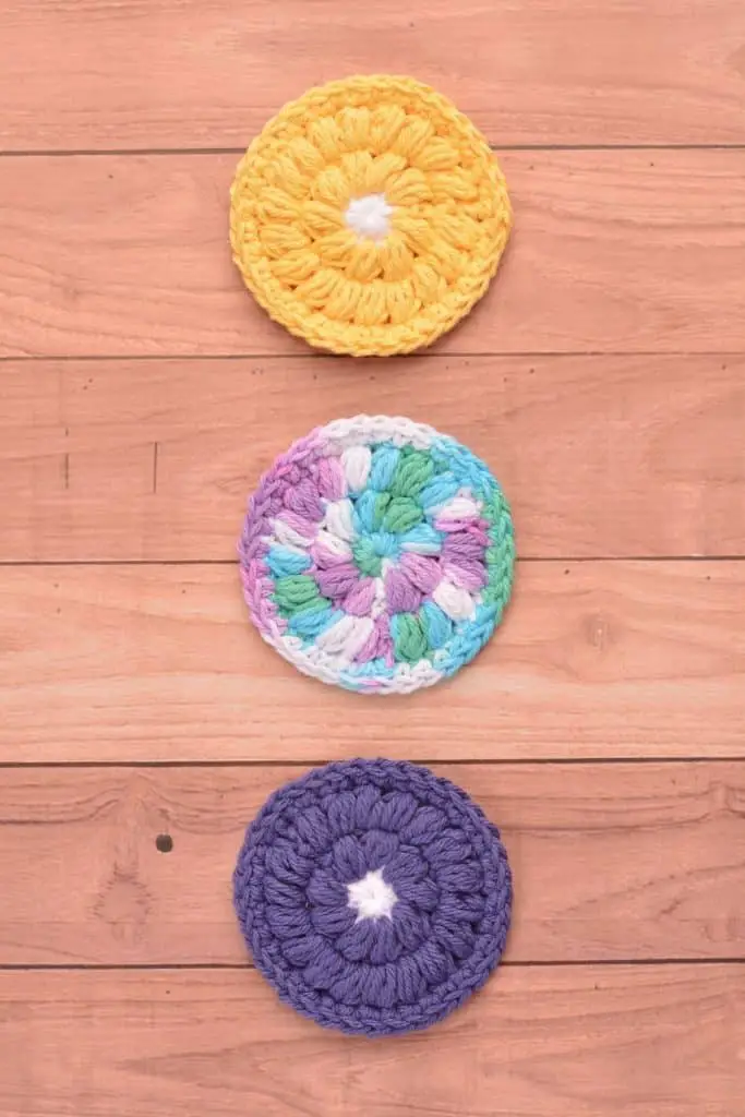 Three face scrubbies on a wooden table. Colors of yellow and dark orchid and ombre yarn.