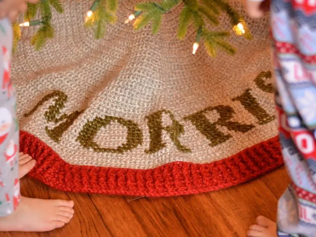 Crochet beginner tree skirt that is grey, with ribbed border in red and lettering in green yarn