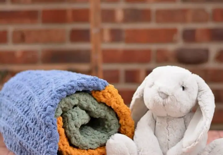 Rolled up denim blue, mustard yellow and dusty sage green baby blanket on a table next to a cuddly grey bunny rabbit
