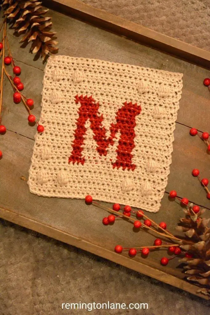 Crochet blanket square in cream yarn with red monogram letter on a wooden tray.