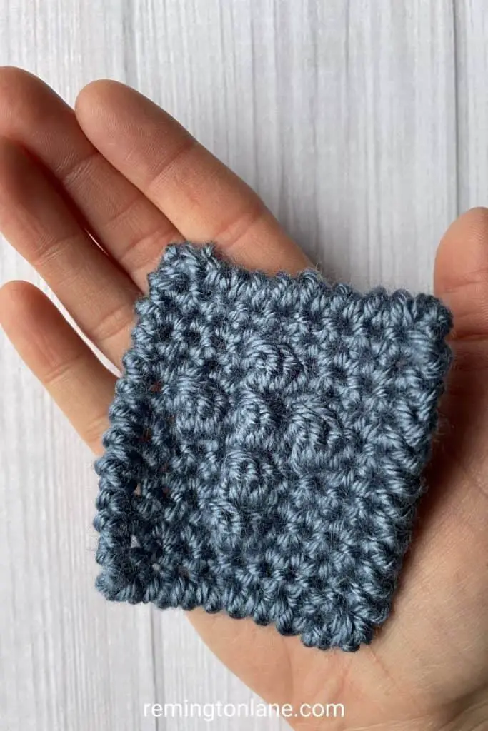 A hand holding a dusty blue crochet prayer square that has a textured cross in the middle