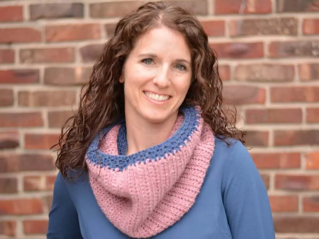 Smiling woman with brown curly hair wearing a handmade cowl in pink yarn with blue trim.