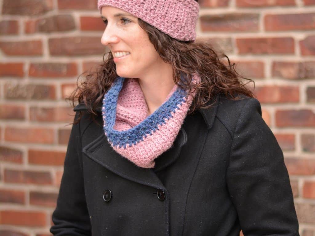 Young woman wearing a black winter coat, styled with a pink crochet ear warmer and pink neck warmer.