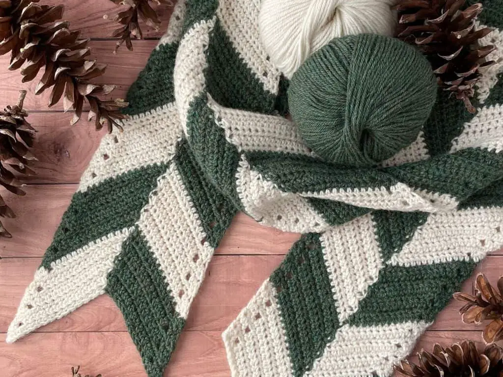 A cream and green scarf with matching yarn balls and pinecones on a wooden table