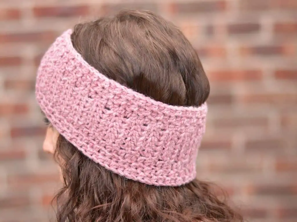 Rear view of a light pink crocheted headband modeled on a young woman with brown curly hair.