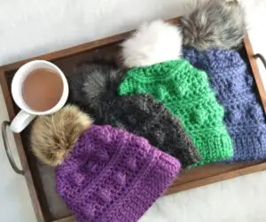 A wooden serving tray filled with a mug of hot chocolate and winter hats crocheted in jewel toned yarn