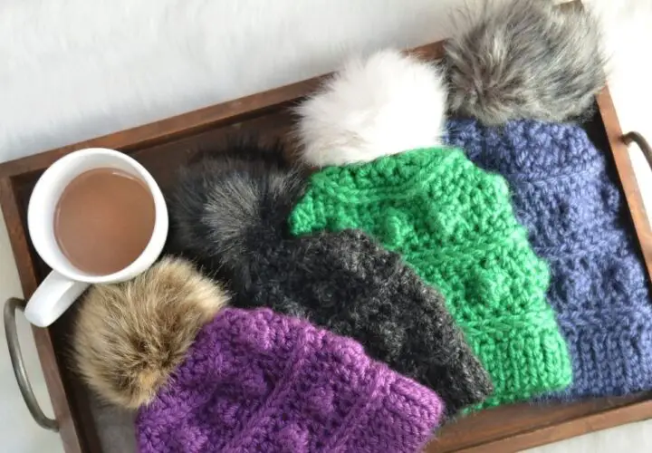 A wooden serving tray filled with a mug of hot chocolate and winter hats crocheted in jewel toned yarn