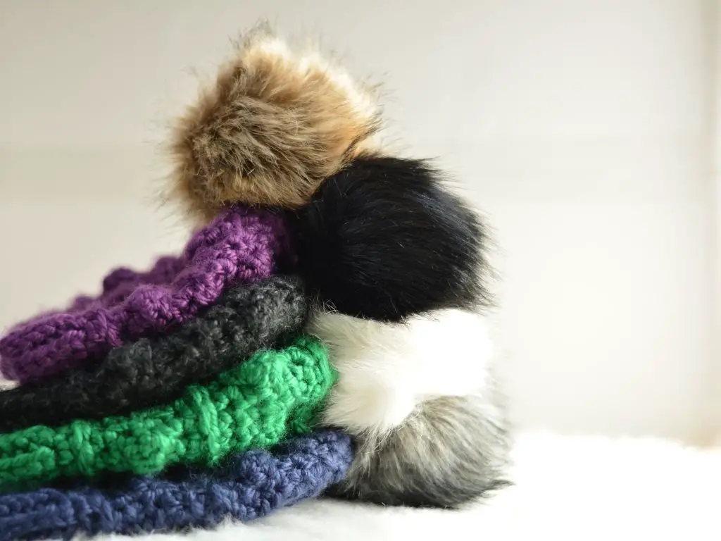 A stack of four jewel-toned winter hats with big faux fur poms on top