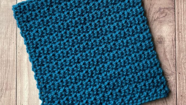 A flat textured square crocheted out of blue yarn on a wooden background