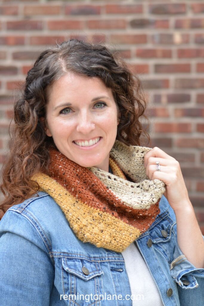 smiling young woman wearing a rustic colored cowl and denim jacket in front of a red brick wall