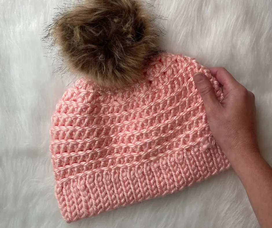 Close up of a light pink crochet hat with a tan pom pom, laying on a piece of white fur