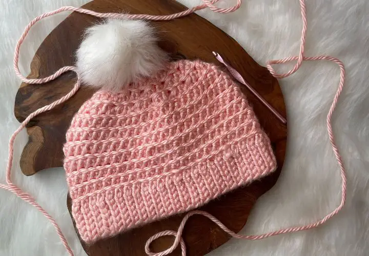 Close up of a crocheted hat in light pink yarn with a white pom pom on top