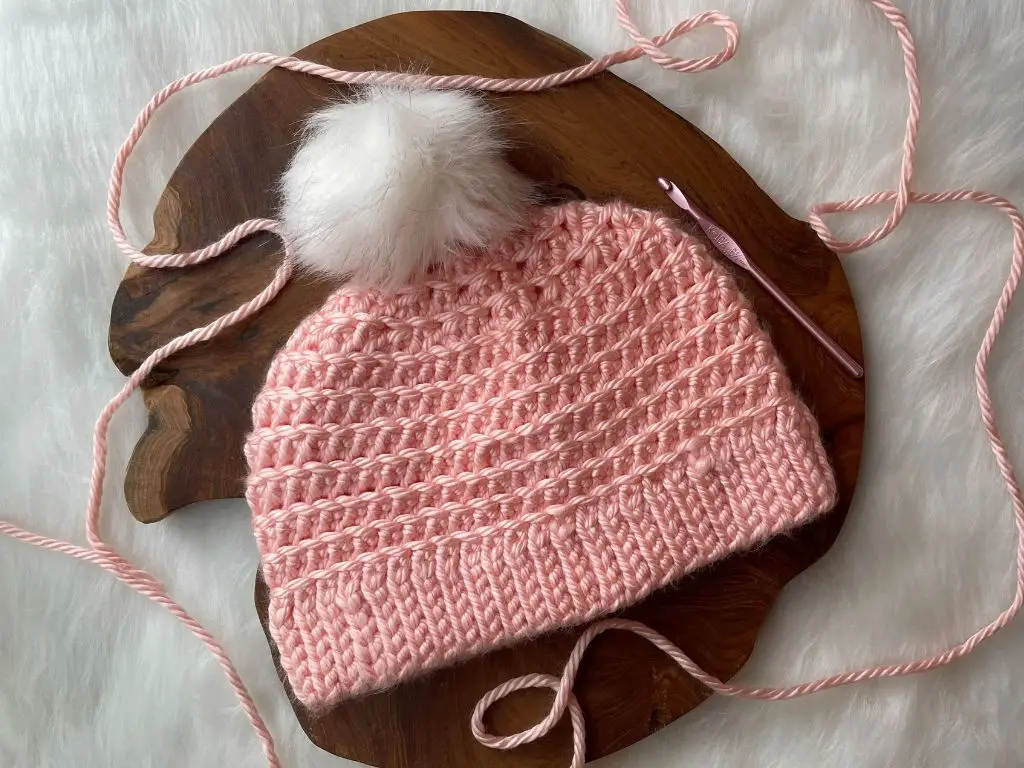 Light pink crochet hat with a white fur pom pom laying on a slab of wood