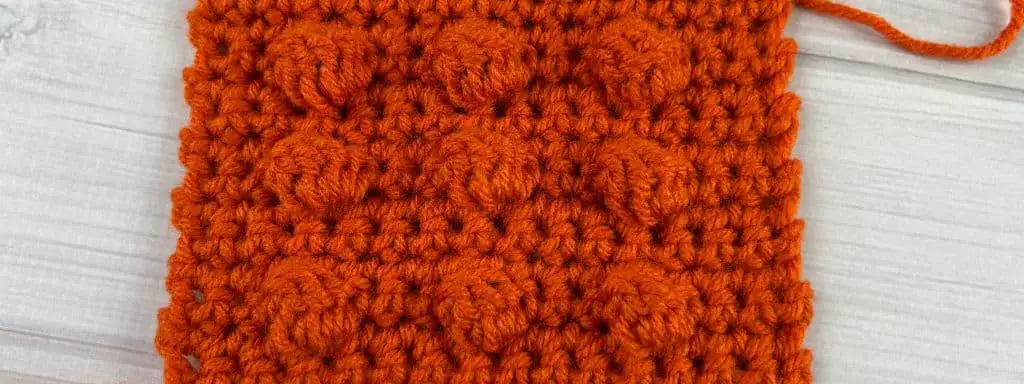 Swatch of fabric made with orange yarn, and has a grid of nine bobbles on it