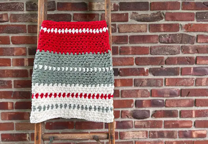 Striped crochet Christmas blanket folded over a wooden ladder leaning against a red brick wall