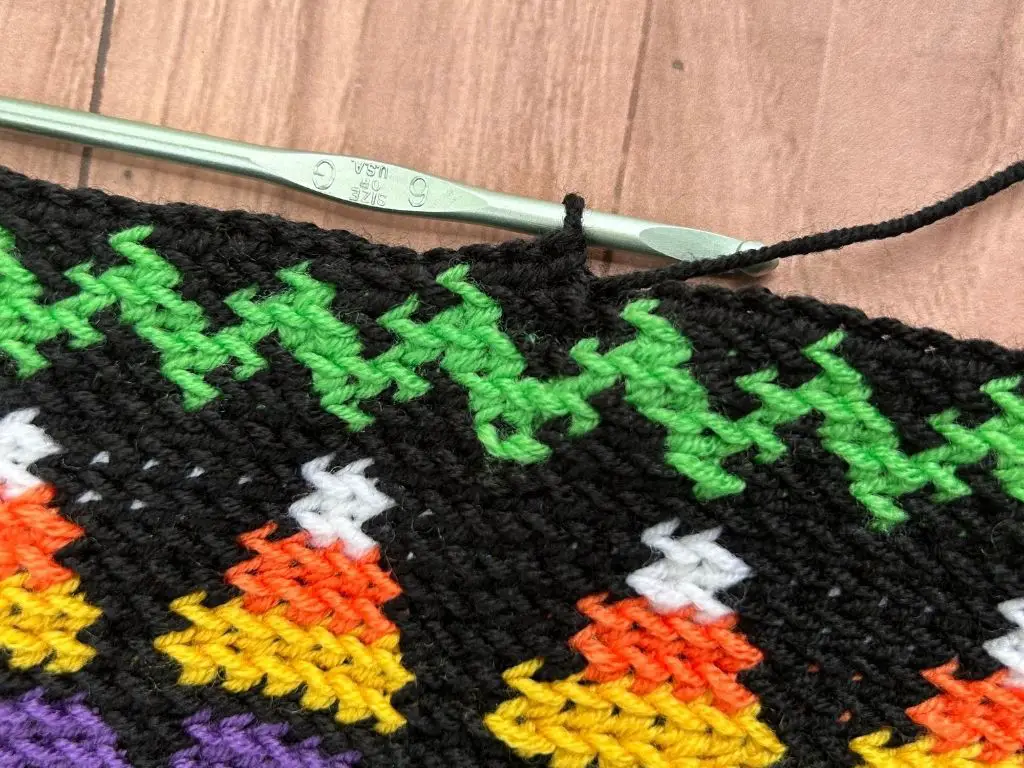 A progress photo of a Halloween-themed cowl laying on a table with the crochet hook working a stitch