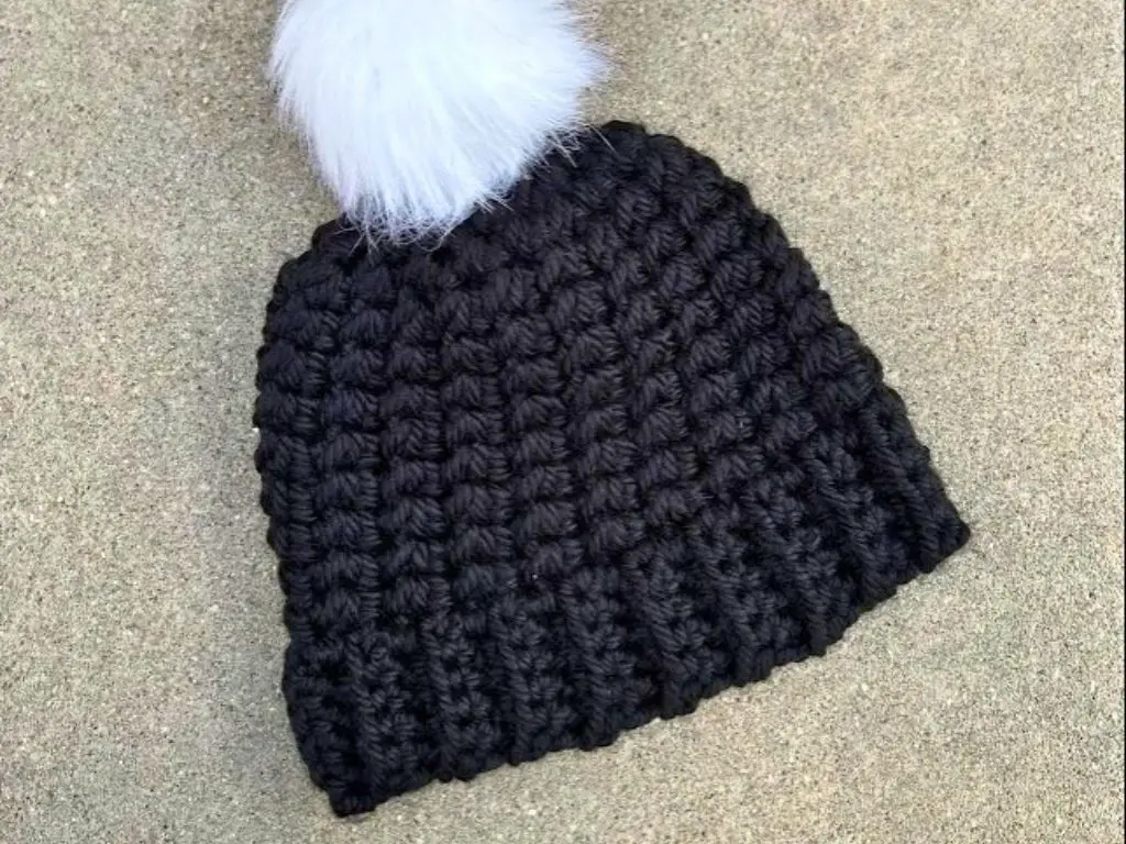 A unisex crochet beanie made with black yarn and a white faux fur pom pom on top.