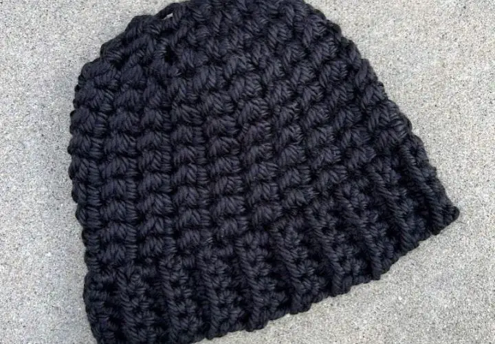 A warm and chunky winter hat crocheted out of black yarn, laying flat on a grey backdrop