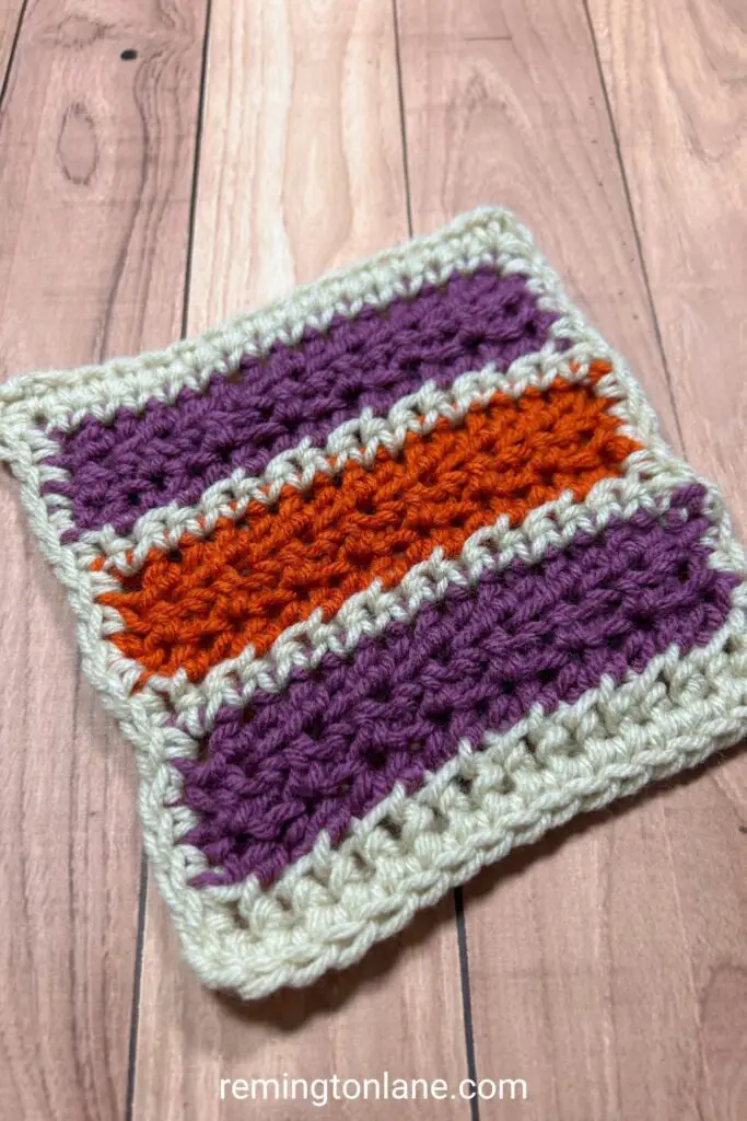 A crocheted blanket square in purple, orange and cream stripes.