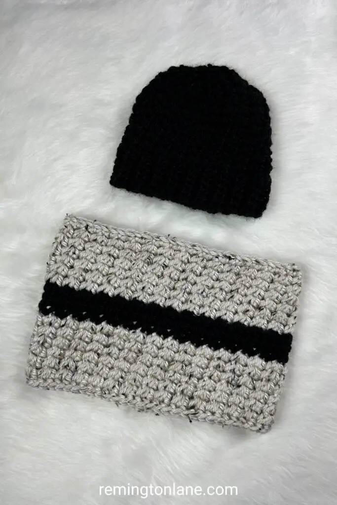 A black crochet hat with coordinating black and grey striped cowl laying on white fur.