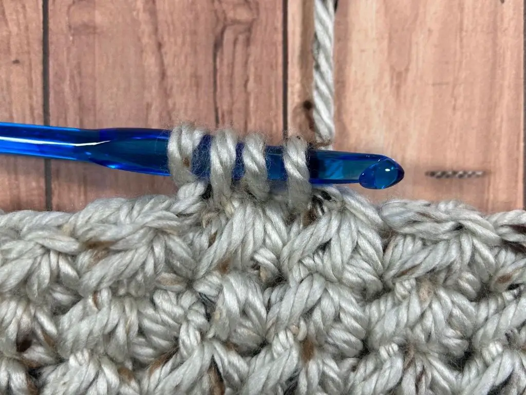 Close-up of grey tweed yarn with a blue plastic crochet hook in the middle of making a trinity stitch.