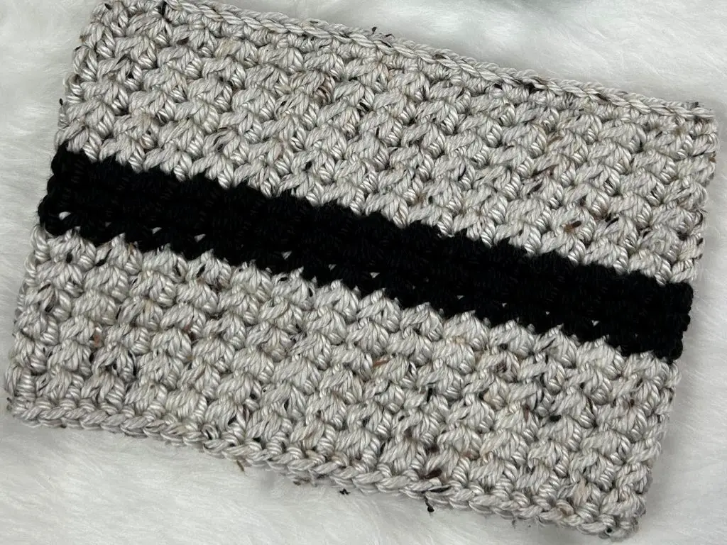 A handmade winter cowl in large stripes of grey tweed and black, laying on a table covered in white fur.