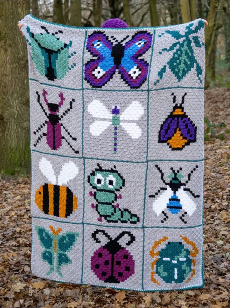 Person in the woods holding up a crochet blanket with 12 different bug images on it
