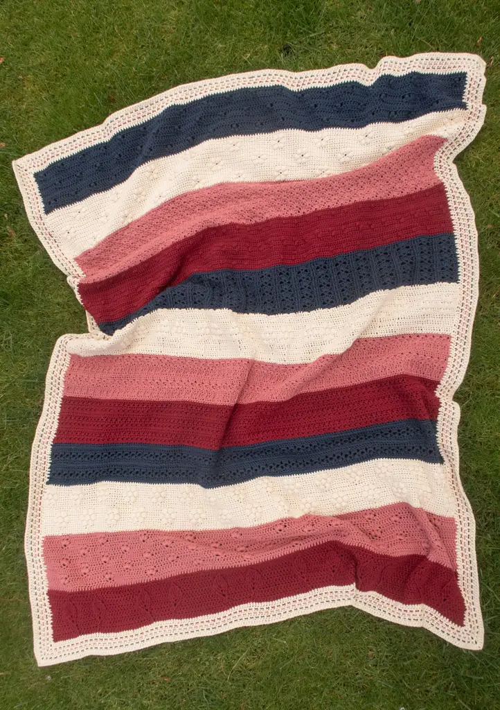 A striped crocheted blanket in navy, red, pink and cream colors, splayed on the grass.