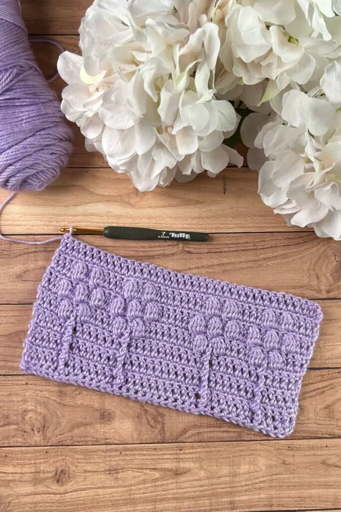 A light purple swatch of a crochet blanket with a flower-inspired texture, next to a skein of yarn and hydrangea flowers.