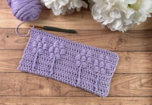 A small swatch of a crochet blanket with a flower-inspired texture, next to a skein of yarn and hydrangea flowers.