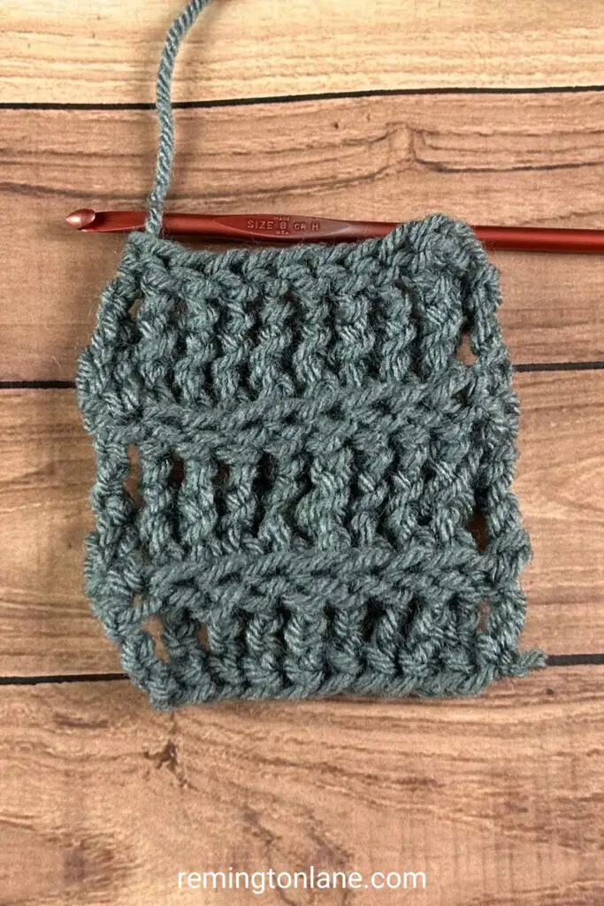 A grey piece of crocheted fabric laying on a wooden table, with a red metal crochet hook ready to make a new row of BPtc stitches.