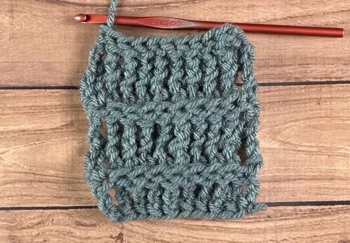 A wooden table with a partially finished swatch of grey yarn showcasing the back post treble crochet stitch after a few rows made.