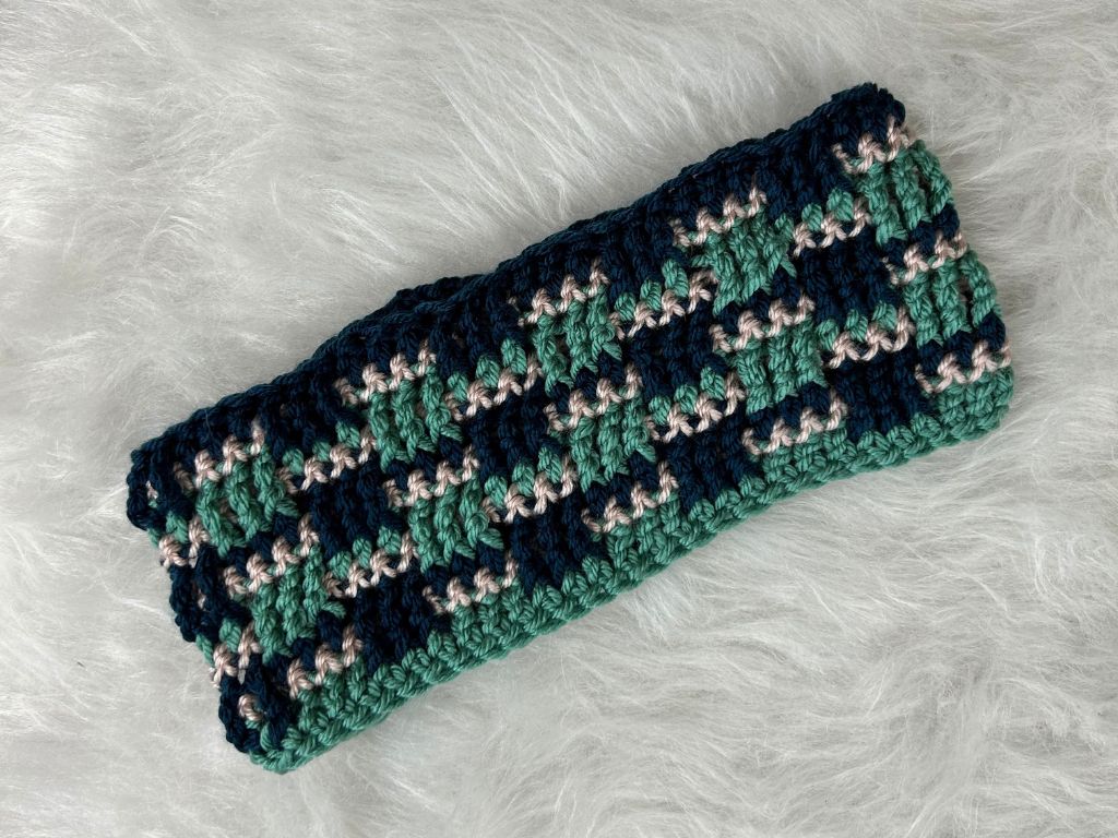 A crocheted ear warmer in navy, blue-green, and light pink stripes laying flat on white fur.