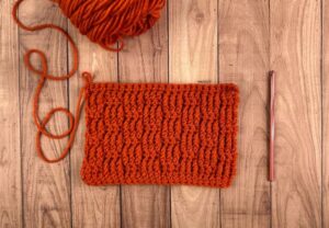 A red-orange yarn crocheted into a small blanket swatch, laying on a wooden background.