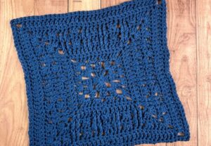 A crocheted blanket square featuring the granny stitch, laying on a wooden backdrop