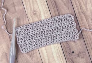 A small strip of textured crocheted fabric with a pearl hook next to it, laying on a table.
